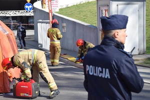 policjant stojący tyłem do zdjęcia oraz strażacy w trakcie działań przed wjazdem do Tauron Areny