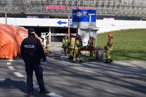 Policjant stojący tyłem do zdjęcia oraz strażacy wynoszący osobę poszkodowaną przed wjzdem do Tauron Areny