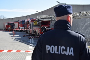 policjnat stojący tyłem do zdjęcia. W tle widać wozy strażackie