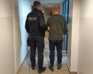 policjant prowadzi zatrzymanego przez korytarz komisariatu. Osoby ustawione tyłem do zdjęcia