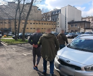 Zatrzymany mężczyzna prowadzony przez funkcjonariuszy przez policyjny parking do radiowozu