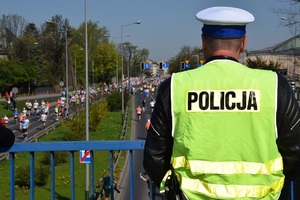 policjant ruchu drogowego stoi na wiadukcie tyłem do obiektywu. Na dole ulicą biegną uczestnicy maratonu