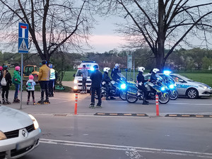 policjanci na motocyklach. Obok znajduję się radiowóz oraz samochód pogotowia ratunkowego. Pojazdy mają włączone sygnały świetlne. Po lewej stronie widać kilka osób biorących udział w wydarzeniu