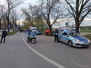 policjant na motocyklu. Obok widać innego funkcjonariusza stojącego obok radiowozu za którym znajduje się pojazd pogotowia ratunkowego z załogą. W tle widać grupę &#039;&#039;rolkarzy&#039;&#039;