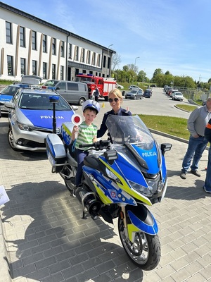 dziecko siedzi na policyjnym motocyklu, obok stoi policjantka w tle radiowozy, wóz strażacki i inne samochody