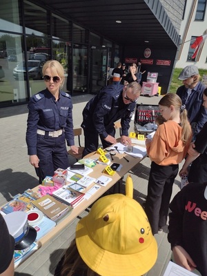 policjant i policjantka przed budynkiem, przy stoisku profilaktycznym, obok uczestnicy wydarzenia