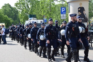 kilkadziesiąt policjantów maszerujących w dwuszeregu przez jedną z krakowskich ulic. Za policjantami widać część radiowozu