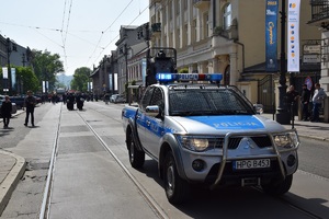 Na pierwszym planie widać policyjny radiowóz. Na drugim planie widać maszerujących policjnatów oraz osoby biorące udział w marszu