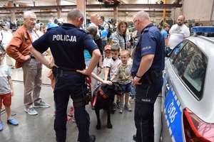 dwaj policjanci z psem służbowym stoją przy radiowozie, obok stoją dzieci i dorośli