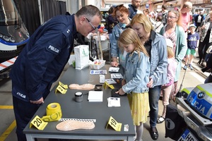policyjny technik kryminalistyki przy stoliku opowiada kobiecie i dziecku o zabezpieczaniu odcisku palców