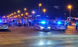 policjanci stojący obok radiowóz z włączonymi sygnałami świetlnymi. Za nimi widać tłum osób. Zdjęcie zrobione w porze nocnej