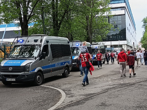 kilka policyjnych radiowozów stojących przed stadionem. Obok widać przemieszczających się kibiców