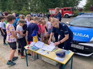 policjantka przy stanowisku profilaktycznym policji z ułożonymi materiałami edukacyjnymi. Obok widać kolejkę dzieci stojących obok policyjnego stanowiska