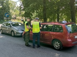 Policjant i funkcjonariusz Straży Granicznej rozmawiają z kierowcą czerwonej taksówki znajdującym się w środku pojazdu. Przed nimi widać innego policjanta rozmawiającego z kierowcą innej taksówki