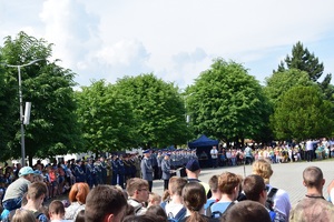 W oddali widać policjantów uczestniczących w uroczystym apelu. Na bliższym planie widać osoby cywilne stojące tyłem do zdjęcia