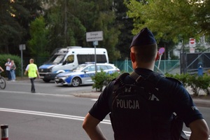 Umundurowany policjant od tyłu, w tle widać radiowozy.