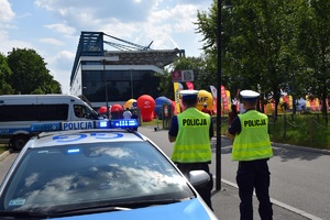 dwóch policjantów ruchu drogowego w kamizelkach odblaskowych obserwujących obiekt sportowy przy ul. Reymonta w Krakowie. Po prawej stronie widać dwa policyjne radiowozy