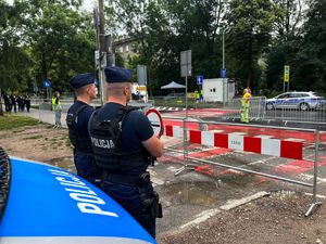 dwóch umundurowanych policjantów stojących przed radiowozem obserwuję otoczenie. W oddali widać policyjny radiowóz. Po lewej stronie w oddali widać patrol straży miejskiej