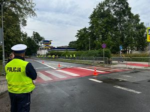 policjant ruchu drogowego stojący przy drodze obserwuje osoby w kamizelkach odblaskowych idące chodnikiem