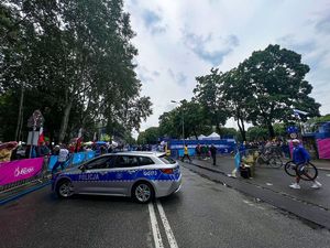 policyjny radiowóz stojący na drodze. W oddali widać zgromadzone osoby. Częśc z nich znajduje się poza wydziloną strefą, druga częśc wewnątrz wydzielonej strefy