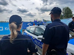 umundurowana policjantka i umundurowany policjant stojący tyłem do zdjęcia przed policyjnym radiowozem