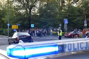 Na pierwszym planie widać policyjne sygnały świetlne. W oddali widać policjanta ruchu drogowego kierującego ruchem oraz pieszych zmierzających na stadion