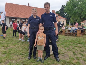 dziecko pozuje do pamiątkowego zdjęcia z policjantką i policjantem, w tle uczestnicy pikniku