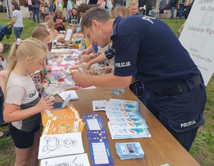 policjant pomaga dziewczynce odbić linie papilarne na pamiątkowej kartce, w tle inni ludzie