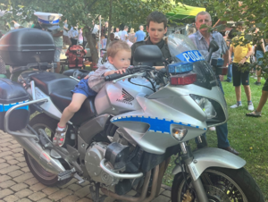 mały chłopiec siedzi na policyjnym motocyklu obok niego stoi starszy chłopiec oraz mężczyzna w tle inni ludzie