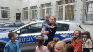 policjantka trzyma dziecko na ręku, obok inne dzieci w tle radiowóz i fragment budynku
