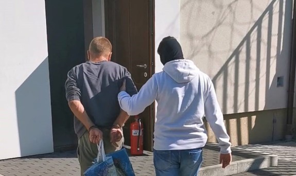 policjant kryminalny prowadzi osobę zatrzymaną z kajdankami na ręce trzymane z tyłu do budynku prokuratury