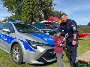 dziewczynka z policyjną tarczą do zatrzymywania pojazdów w ręce, pozuje do zdjęcia z policjantem na tle radiowozu