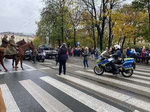 policjant na motocyku znajdujący się w rejonie przejścia dla pieszych. Na zdjęciu widać także dwóch mężczyzn w umundurowaniu na koniach, pojazdy wojaska oraz zgromadzoną widownię