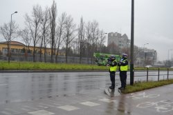 dwóch policjantów ruchu drogowego, jeden trzyma w ręku ręczny miernik prędkości tzw. radar oraz tarczę do zatrzymywania pojazdów. W tle jezdnia i jadące pojazdy, pas zieleni oraz budynki.