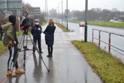 policjantka ruchu drogowego udziela wywiadu telewizyjnego. W tle jezdnia, poruszające się pojazdy oraz pas zieleni.